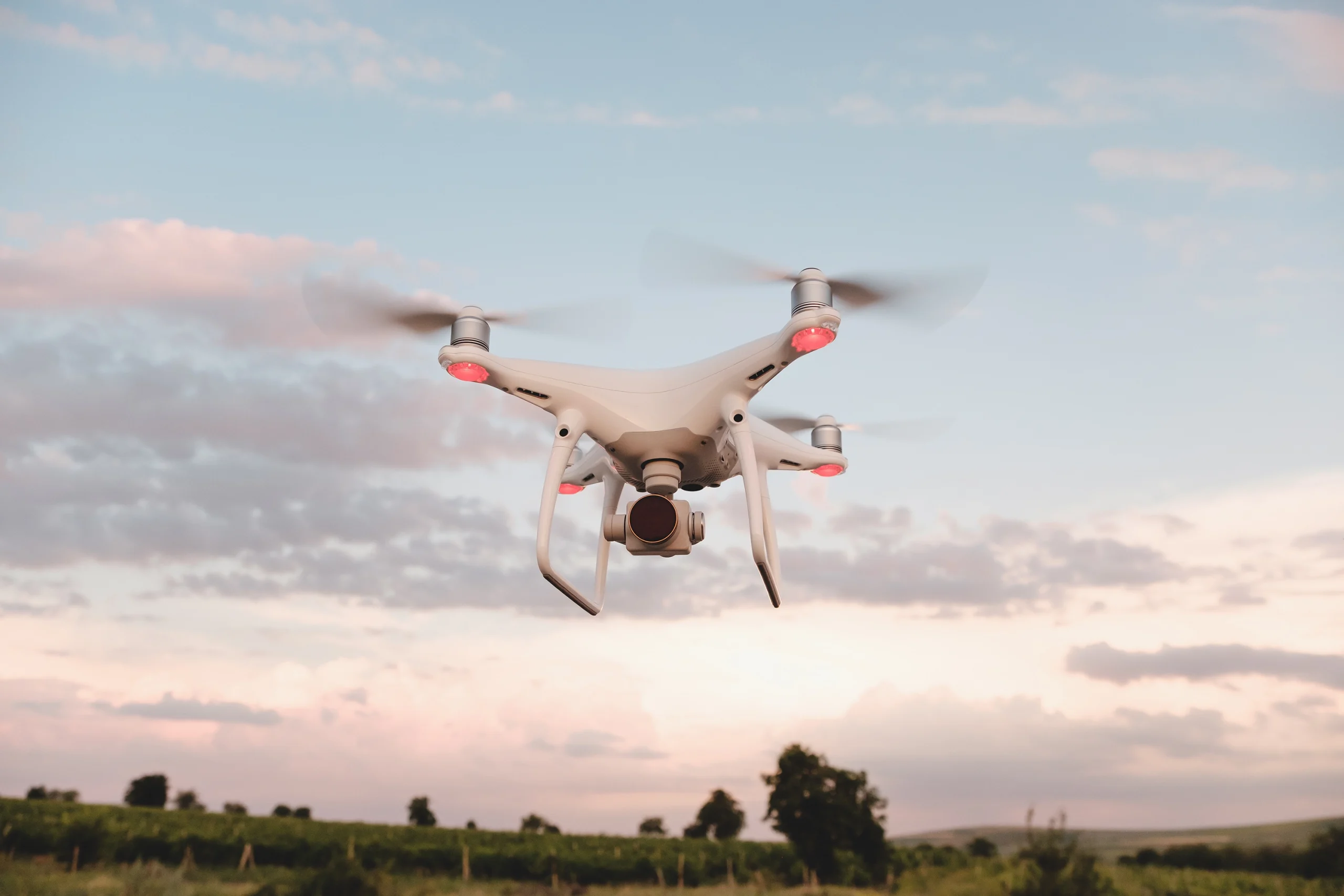 White security drone flying above dark trees with pink and blue colored sky in background